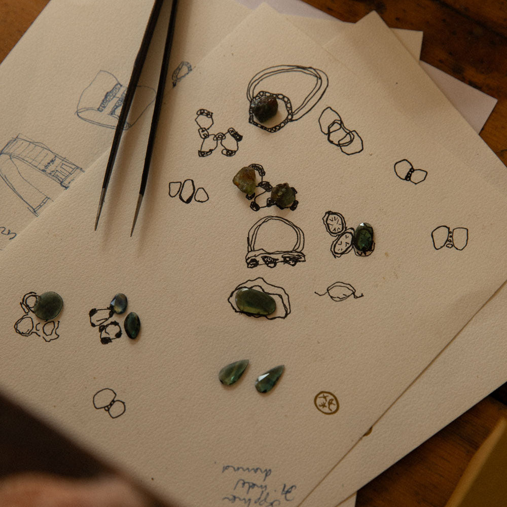 A close-up of sketchbooks and papers on a wooden table, featuring hand-drawn jewellery designs, small loose green gemstones, and tweezers.
